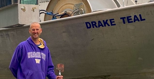 Andy Mitby of Drake Teal Fisheries stands in front of his boat in a University of Washington sweatshirt.