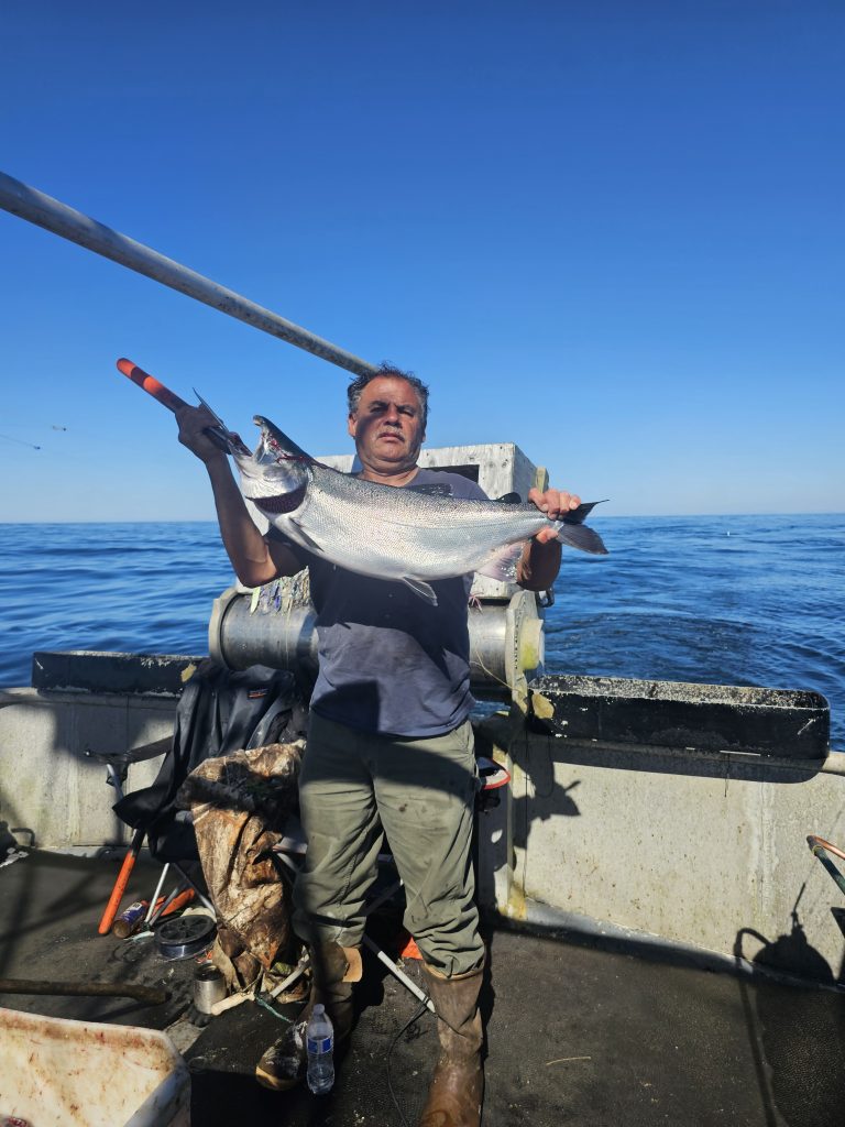 Roger Bain stands on his fishing vessel showing off a large salmon with water to his back.