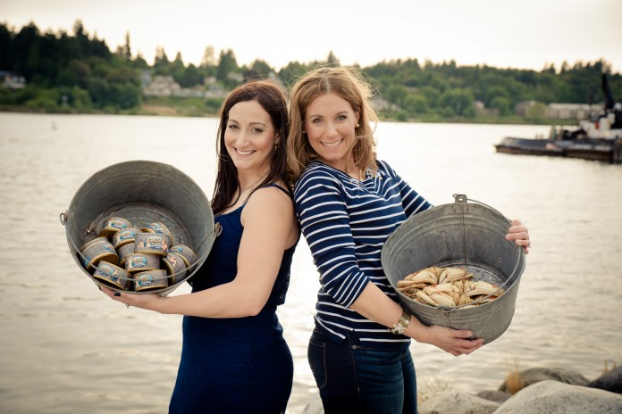 Libie Cain and Teresa Reeves from Sassy Seafood pose back to back, holding two buckets with canned albacore tuna and Dungeness crab.